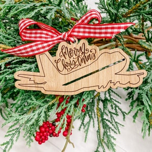 May include: Wooden airplane ornament with the text "Merry Christmas" in cursive. The ornament is decorated with a red and white gingham ribbon.