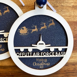 May include: White wooden Christmas ornament with a blue background and gold glitter. The ornament features a silhouette of a plane with the text "Offutt Air Force Base" and "Merry Christmas 2021". There are also gold reindeer and Santa Claus in a sleigh.