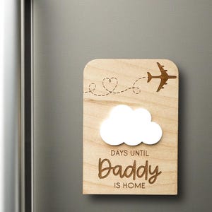 May include: Wooden plaque with a cloud, airplane, and the text "DAYS UNTIL Daddy IS HOME." The plaque has a light wood grain finish and is designed to be a countdown to a special event.