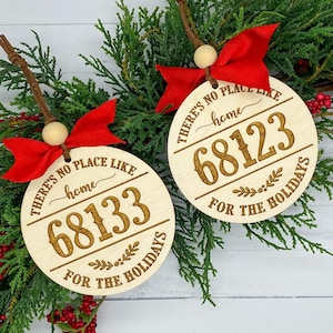 May include: Two round wooden ornaments with red ribbon and a wood bead. The ornaments are engraved with the text "There's no place like home" and a five-digit number. The text "For the holidays" is also engraved on the ornaments.