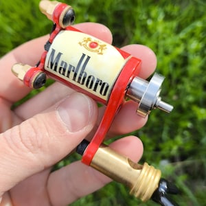 May include: A custom tattoo machine with a red frame and gold accents. The machine's barrel is white with the "Marlboro" logo printed on it. The machine is held against a blurred green background.