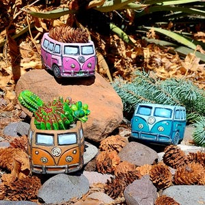 May include: Three colorful ceramic planters shaped like vintage vans. The planters are painted in shades of pink, orange, and blue, with details like headlights and windows. Each planter holds a different succulent or dried plant arrangement, creating a unique decorative display.