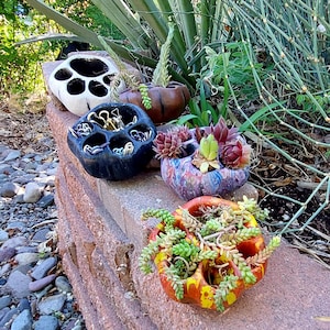 May include: A collection of handmade succulent planters in various colors and designs. The planters are made of ceramic and feature multiple openings for planting. The succulents are a mix of green, red, and purple. The planters are arranged on a brick wall.