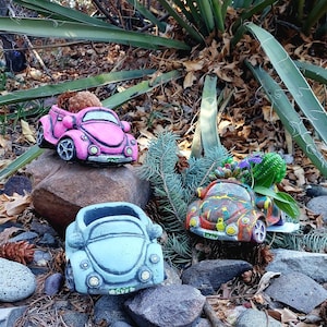 May include: Three colorful, decorative car-shaped planters. One is light blue, one is pink, and the other is multi-colored. Each planter has a detailed design and is filled with plants. The planters are sitting on rocks.