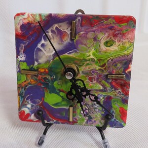 May include: Square clock with a colorful abstract design in shades of purple, green, red, and white. The clock has black hands and brass-colored accents. It is mounted on a black metal stand.