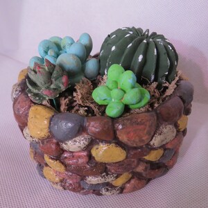 May include: A small, round, stone planter with a variety of succulents. The planter is made to look like a stone wall with different colored stones. The succulents are green, blue, and white.