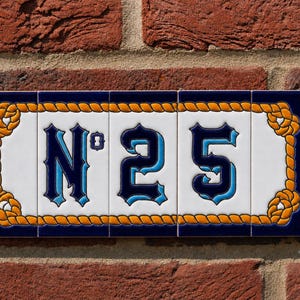 May include: A rectangular house number sign composed of five white ceramic tiles featuring the text N°25 in a dark blue and light blue font. The design is framed by a decorative orange rope border set against a deep navy blue background.
