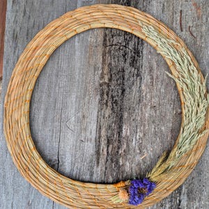 May include: A circular wreath made of woven straw, accented with dried wheat and purple flowers. The wreath is a natural tan color with orange thread accents, perfect for rustic home decor.