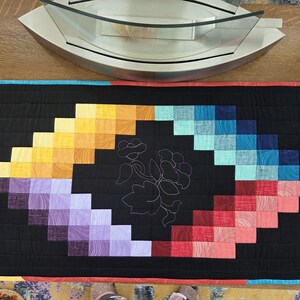 May include: A rectangular quilt with a black background and a central design of a flower. The design is surrounded by a rainbow of colors, including yellow, orange, purple, blue, and red. The quilt has a yellow border.