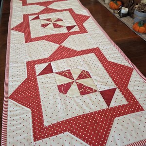 May include: A rectangular quilted table runner with a repeating pattern of red and white geometric shapes. The runner features a white background with red polka dots and red star and square designs. The edges are trimmed with red and white stripes.