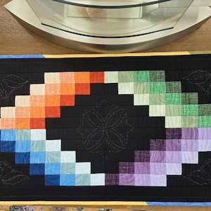May include: A colorful, handmade quilt with a black background. The quilt features a central design of a stylized flower and a gradient of colors, including blue, orange, green, and purple. The quilt is rectangular and has a black border.