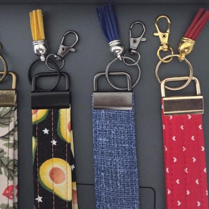 May include: A collection of keychains with fabric wrist straps and colorful tassels. The straps feature various patterns, including floral, avocado, denim, red hearts, and a celestial design. Each keychain has a metal clasp and ring.