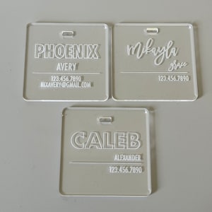May include: Three clear acrylic name tags with white lettering. The top tag reads "PHOENIX AVERY 123.456.7890 NIXAVERY@GMAIL.COM". The middle tag reads "Mikayla Grace 123.456.7890". The bottom tag reads "CALEB ALEXANDER 123.456.7890".