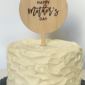 May include: A white cake with a wooden cake topper that says "Happy Mother's Day" in black lettering.