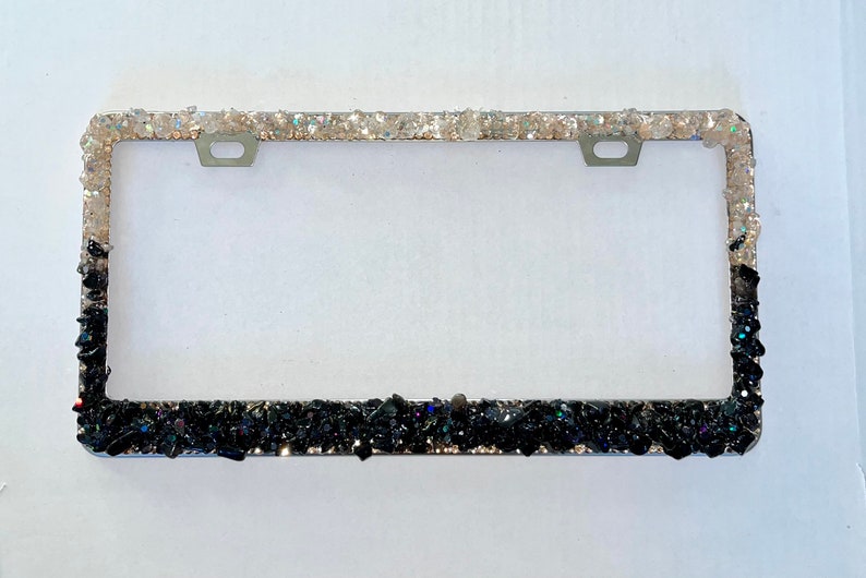 May include: A silver license plate frame with a black and white glitter design. The top half of the frame is covered in white glitter, while the bottom half is covered in black glitter.