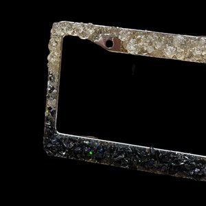 May include: A silver license plate frame with a black and white glitter design. The top half of the frame is covered in white glitter, while the bottom half is covered in black glitter.