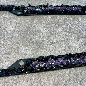 May include: Black license plate frames adorned with purple and iridescent crystals and black stones. The frames have a sleek, modern design, with a row of sparkling embellishments.