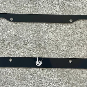 May include: Black license plate frame with a white skeleton hand making the rock and roll hand gesture.