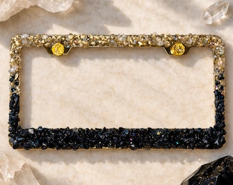 May include: A rectangular license plate frame featuring a gradient design of small, textured stones. The top section is covered in clear, light-reflecting crystals, while the bottom section transitions into deep black, dark-toned stones.