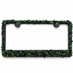 May include: A rectangular license plate frame featuring a dense, textured surface covered in small, irregular dark green and black stone chips. The frame has two circular mounting holes at the top, each accented with a matching stone-covered cap.