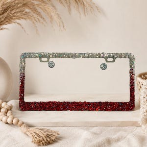 May include: A license plate frame with a gradient of red glitter at the bottom, transitioning to silver glitter at the top. The frame is adorned with sparkling embellishments and has two silver mounting holes.