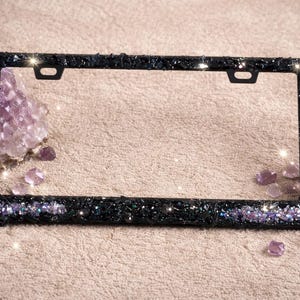 May include: A black license plate frame encrusted with dark glitter and purple iridescent accents. The frame is surrounded by amethyst crystals on a neutral surface. The frame has two mounting holes at the top.