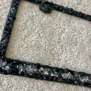 May include: A rectangular license plate frame decorated with a dense arrangement of black, polished stones and small, clear, reflective crystal-like fragments embedded along the bottom edge, creating a textured, dark, and shimmering appearance.
