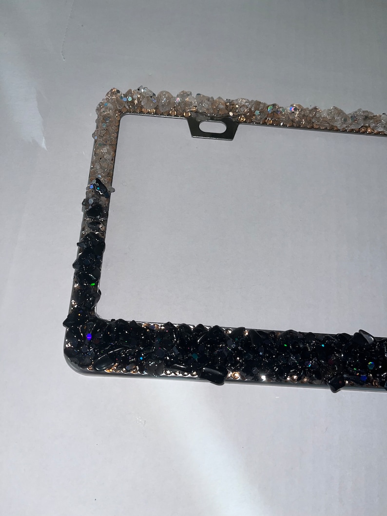 May include: A black and clear crystal embellished license plate frame. The frame is made of metal and has a black finish. The crystals are arranged in a random pattern around the frame.