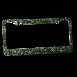 May include: A license plate frame with a silver border, encrusted with small, irregularly shaped green stones. The stones are tightly packed together, creating a textured surface. The frame is rectangular with rounded corners, set against a black background.