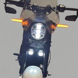 Può includere: Una moto nera con parafango anteriore bianco e parabrezza trasparente. La moto ha un faro bianco brillante e due luci bianche più piccole sotto. Gli indicatori di direzione arancioni sono sul manubrio.