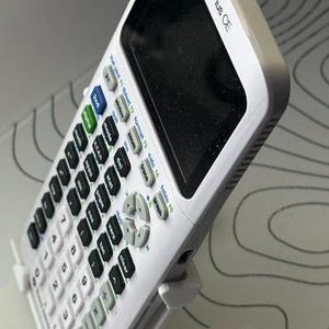 May include: A white TI-84 Plus CE graphing calculator with a black screen and many buttons. The calculator is supported by a white stand, angled towards the viewer. The calculator has the text "TI-84 Plus CE" printed on the top.
