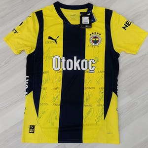 Könnte beinhalten: Ein gelb-dunkelblau gestreiftes Kurzarmtrikot mit dem Wappen des Fenerbahçe Spor Kulübü, einem Puma-Logo und dem in Weiß gedruckten Schriftzug Otokoc. Die Vorderseite des Trikots ist mit zahlreichen schwarzen Tintenunterschriften versehen und hat ein Etikett.