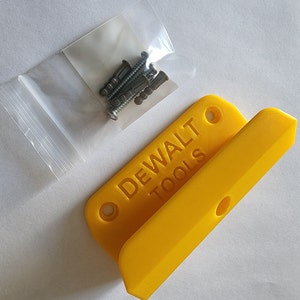 Puede incluir: Soporte de pared de plástico amarillo con el texto "DEWALT TOOLS" y dos agujeros. Se incluye una pequeña bolsa de tornillos.