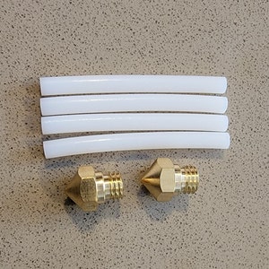 May include: A top-down view of 3D printer components. The image shows four white, cylindrical tubes and two brass nozzles with threaded bases. The items are arranged on a speckled, light brown surface.
