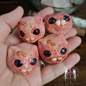 Könnte beinhalten: Vier rosa, runde, kreaturenartige Skulpturen mit schwarzen Augen und kleinen, weißen Zähnen. Die Skulpturen haben eine strukturierte Oberfläche mit kleinen, dunklen Punkten und ohrartigen Vorsprüngen. Die Objekte werden in einer Hand gehalten.
