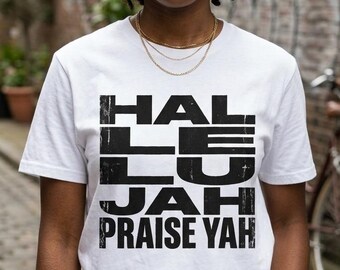 May include: White t-shirt with the text "HALLELUJAH PRAISE YAH" in a bold, black, distressed font. The shirt is worn by a person with dark hair and gold jewelry, standing in an outdoor setting.