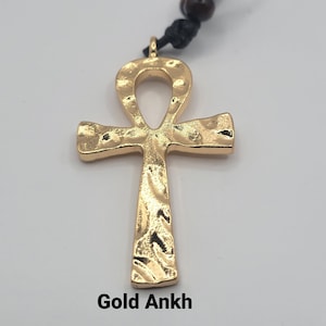 May include: A gold Ankh pendant with a textured surface, hanging from a black cord with a brown bead. The pendant is a symbol of life and eternal life in ancient Egyptian culture.