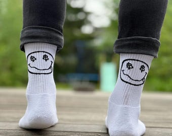 Op de afbeelding: Witte geribbelde sokken met een zwart smiley-gezicht ontwerp, met dollartekens voor ogen. De sokken worden gedragen met een donkergrijze broek. De afbeelding is genomen vanuit een lage hoek, met de sokken en de onderbenen.