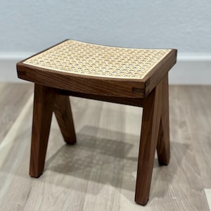 Puede incluir: Un pequeño taburete cuadrado de madera con un asiento de ratán tejido. El taburete tiene cuatro patas anguladas y está hecho de madera de color marrón oscuro. El asiento es de color beige claro con un patrón repetitivo. El taburete está diseñado para sentarse.