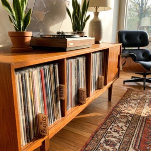 May include: A mid-century modern wooden record cabinet with a turntable on top. The cabinet holds numerous vinyl records, with dividers labeled "The Beatles" and other bands. Two potted plants sit on top.