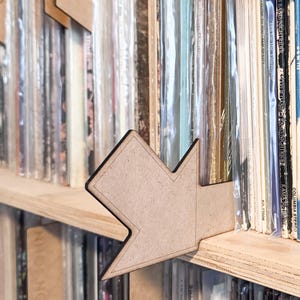May include: A wooden arrow-shaped shelf divider with a black outline, placed on a wooden shelf among vinyl records. The records are in protective sleeves. The shelf divider is made of light brown wood.