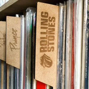 May include: A close-up of a shelf with vinyl records organized by artist. The shelf has wooden dividers with the names "Rolling Stones", "Prince", "Jimi Hendrix", and "Aerosmith" engraved on them.