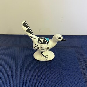 May include: A white ceramic roadrunner figurine with black and blue geometric designs. The bird has a long tail and is perched on a small, round base. The figurine is painted with black and blue accents.