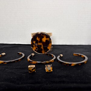 May include: A jewelry set featuring a wide cuff bracelet with a large circular tortoise shell pattern inlay, three thin open-ended bangles with a matching tortoise shell design, and two small square stud earrings with a tortoise shell finish.