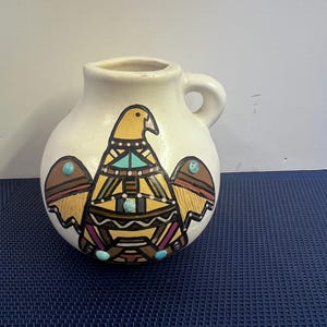 May include: A small, off-white ceramic vase with a handle. The vase features a colorful, geometric design of a bird with turquoise accents. The bird design is centered on the front of the vase, with brown and yellow wings.
