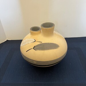 May include: A tan ceramic vase with two spouts and a round body. The vase is decorated with gray and black feather designs. The vase has a gray rim and a black stripe around the base.