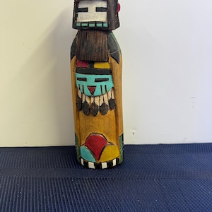 May include: A hand-carved wooden Native American kachina doll. The doll features a detailed face with a dark brown wig, a turquoise face, and a tan body. The doll is painted with red, yellow, and black accents. The base is black and white.