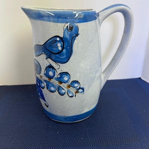 May include: A ceramic pitcher with a blue bird design. The pitcher has a white base with blue accents on the rim, handle, and base. The bird is painted in a bold blue color, perched on a branch with blue berries. The pitcher is approximately 7 inches tall.