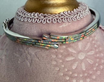 May include: A silver-toned hinged bangle bracelet featuring a multi-layered design with inlaid opal segments. The inlay displays a vibrant play of color with flashes of orange, green, and white against a metallic frame with a secure clasp.