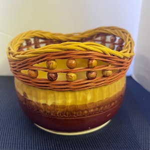 May include: A decorative bowl with a woven rim and a ceramic base. The rim is made of woven yellow and brown material, and the base is a mix of yellow and brown with a textured pattern. Wooden beads are incorporated into the design.